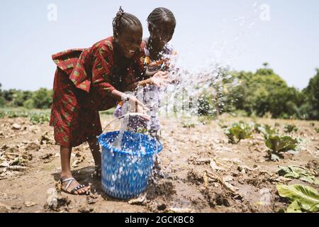 Zwei verspielte kleine afrikanische Schwestern bewässern das Kohlfeld ihres Vaters, mit geschlossenen Augen und einem breiten Lächeln im Gesicht Stockfoto