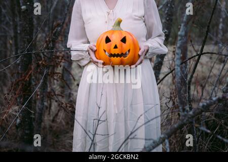 Weibchen in schwarzem Kleid steht an Halloween mit glühender Kürbis-Laterne in dunklem Holz und schaut in die Kamera Stockfoto