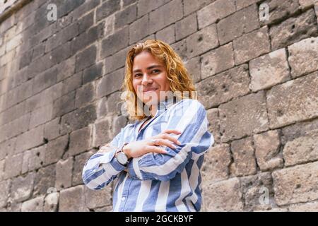 Von unten begeistert Männchen mit langen Haaren stehend mit gekreuzten Armen in der Nähe Steinmauer in der Straße und Blick auf die Kamera Stockfoto