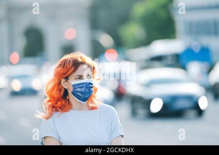 Weibchen mit roten Haaren auf schützender medizinischer Maske in der Stadtstraße während der Coronavirus-Epidemie Stockfoto
