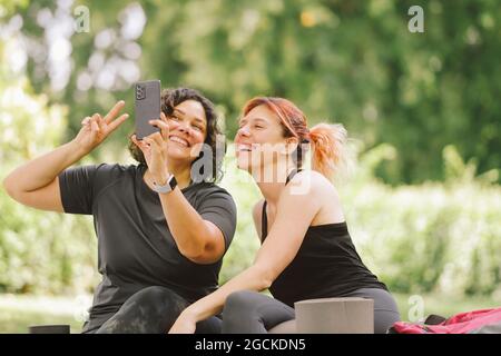 Fröhliche junge multirassische Freundinnen in lässiger Kleidung, die lächeln und eine Siegesgeste zeigen, während sie Selfie auf dem Smartphone machen, das sich in grüner Kleidung ausruht Stockfoto