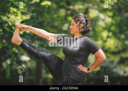 Eine dicke Hündin mit der Hand an der Taille, die in Utthita Hasta Padanguschthasana steht, während sie Yoga praktiziert und im Park nach vorne blickt Stockfoto