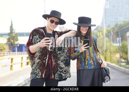 Entzücktes Paar in trendigen Hipster-Kleidern, die im Sommer in der Stadt stehen und Mobiltelefone benutzen Stockfoto