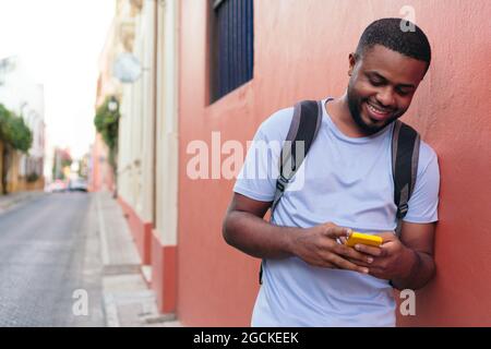 Mann mit Rucksack überprüft Handy, während er sich an die Wand lehnt Stockfoto