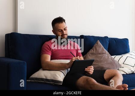 Vollblut selbstbewusster junger bärtiger, ethnischer Freiberufler in legerer Kleidung und Kopfhörern, die auf einem bequemen Sofa sitzen und aus der Ferne auf Ta arbeiten Stockfoto