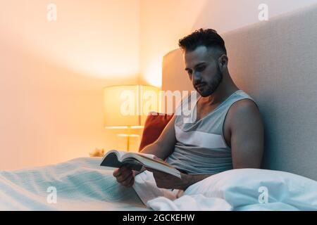 Nachdenklicher Mann, der unter der Decke auf dem Bett saß und vor dem Schlafen interessante Geschichten in einem Buch las Stockfoto