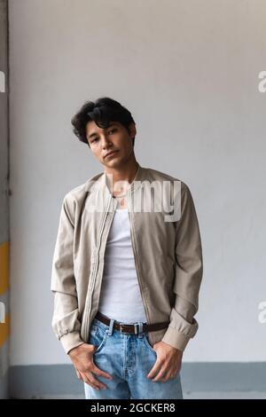 Porträt eines jungen lateinischen Mannes in legerer Kleidung, der selbstbewusst auf die Kamera blickt, mit den Händen auf den Taschen, während er auf dem Parkplatz steht Stockfoto