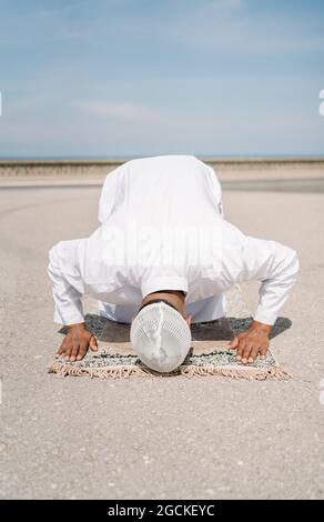 Nicht erkennbarer muslimischer Mann, der auf dem Teppich kniet und den Boden mit der Stirn berührt, während er an sonnigen Tagen am Sandstrand betet Stockfoto