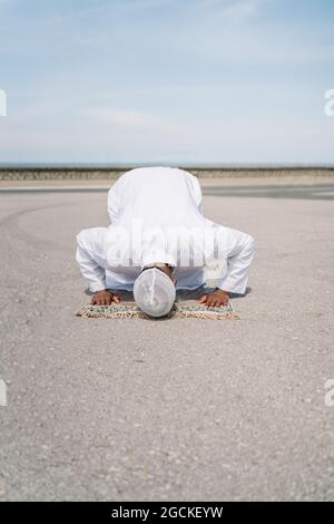 Nicht erkennbarer muslimischer Mann, der auf dem Teppich kniet und den Boden mit der Stirn berührt, während er an sonnigen Tagen am Sandstrand betet Stockfoto