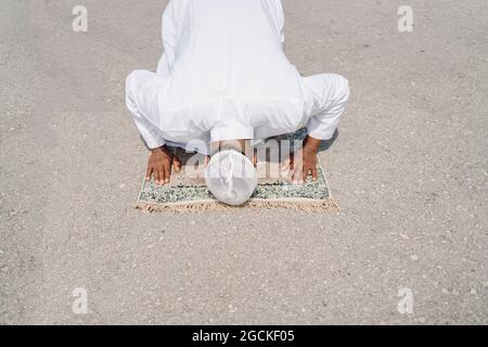 Nicht erkennbarer muslimischer Mann, der auf dem Teppich kniet und den Boden mit der Stirn berührt, während er an sonnigen Tagen am Sandstrand betet Stockfoto