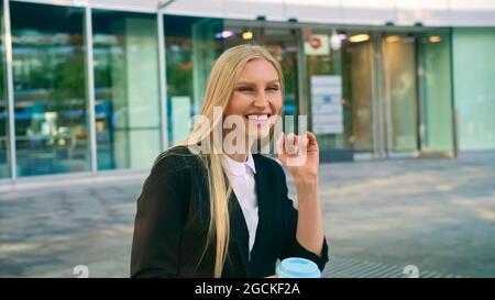 Elegante, moderne Frau, die auf der Bank sitzt und mit der Hand zum Freund winkt, während sie auf dem Smartphone draußen spricht Stockfoto