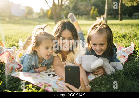 Glückliche junge Frau und niedliche kleine Töchter liegen auf der Decke und machen Selfie auf dem Smartphone, während sie gemeinsam auf der grünen Wiese im Sommerpark Spaß haben Stockfoto