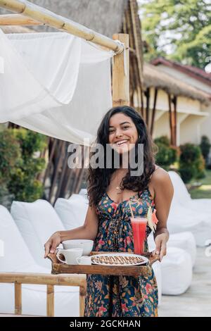 Lächelnde weibliche Touristin in der Tray mit leckerem Frühstück, während sie während der Reise auf die Kamera schaute Stockfoto