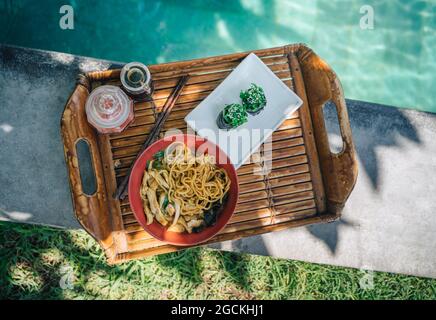 Blick von oben auf das Tablett mit Gunkan-Sushi-Brötchen mit chuka gegen eine Schüssel gekochter Pasta und Hähnchenstückchen am Pool Stockfoto