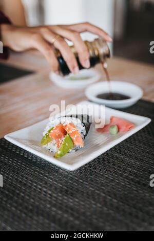 Crop unkenntlich weiblich Gießen Sojabohnensauce in Schüssel gegen leckere Sushi-Kegel mit Lachs und Avocado Scheiben auf dem Teller Stockfoto