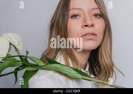 Ruhiges junges Weibchen mit zart blühender weißer Madonna Lily Blume, die im Studio die Kamera anschaut Stockfoto