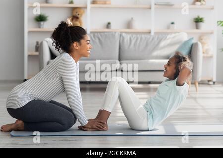 Schwarze Mutter und Tochter in der Sportbekleidung trainieren gemeinsam zu Hause, sitzen auf Fitness-Matte und arbeiten an Bauchmuskeln, tun knirscht, Seite wetteifern Stockfoto