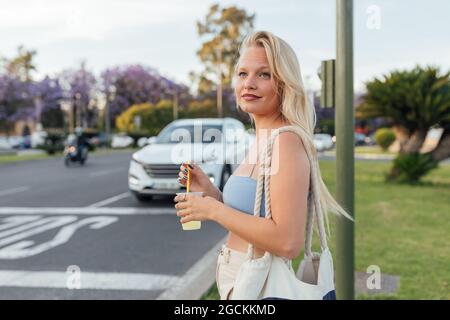 Seitenansicht einer fröhlichen Frau, die im Sommer auf der Straße mit kalter Limonade in einem Plastikbecher steht Stockfoto