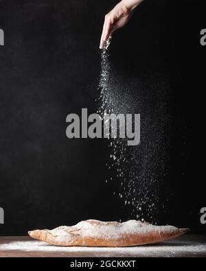 Ernte anonymen Bäcker, der weißes Mehl auf frisch gebackenem handwerklichem Baguette-Brot vor schwarzem Hintergrund streut Stockfoto