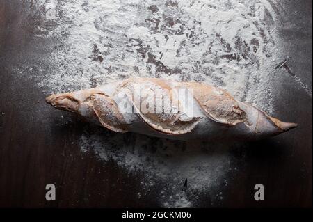 Appetitlich frisch gebackenes Baguette mit knuspriger Kruste auf Holztisch mit weißem Mehl vor schwarzem Hintergrund gelegt Stockfoto