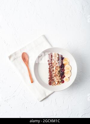 Draufsicht auf eine Schüssel mit weißem Joghurt und verschiedenen Cerealien und Bananenscheiben, serviert für ein gesundes Frühstück auf dem Tisch mit Holzlöffel Stockfoto
