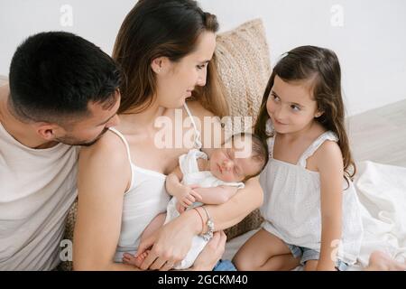 Nettes kleines Mädchen, das auf der Couch sitzt, mit lächelnden, glücklichen jungen Eltern, die das neugeborene Baby halten Stockfoto