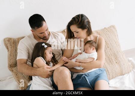 Nettes kleines Mädchen, das auf der Couch sitzt, mit lächelnden, glücklichen jungen Eltern, die das neugeborene Baby halten Stockfoto