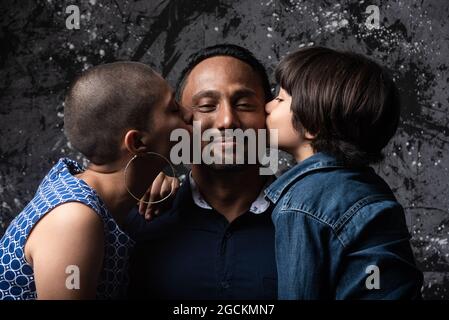 Multiethnisch liebende Frau und Teenager-Sohn küssen Mann auf Wange auf dunklem Hintergrund im Studio Stockfoto