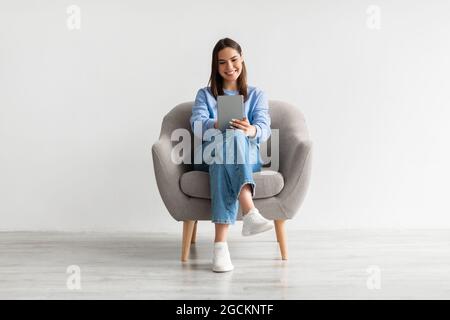 Porträt einer lächelnden jungen Frau mit digitalem Tablet, das im Sessel sitzt und online gegen die weiße Studiowand arbeitet Stockfoto