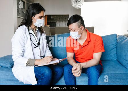 Fröhliche weibliche Praktizierende im medizinischen Kleid im Gespräch mit Teenager-Jungen mit Down-Syndrom und Notizen auf Zwischenablage während der Verabredung zu Hause Stockfoto