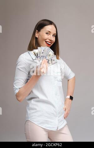 Ein Bild einer lustigen jungen Dame in einem weißen Sommerhemd hält Geld und blickt auf einem grauen Hintergrund zur Kamera Stockfoto