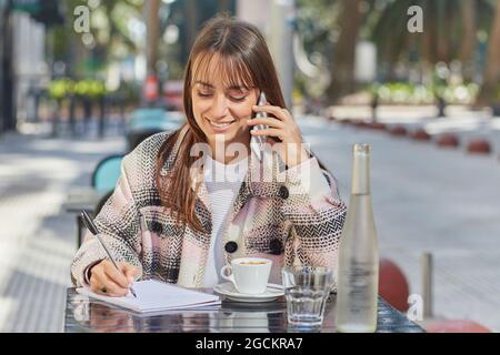 Positive junge Frau, die sich während des Telefongesprächs Notizen im Notizbuch gemacht hat, während sie im Café im Freien in der Stadt am Tisch sitzt Stockfoto