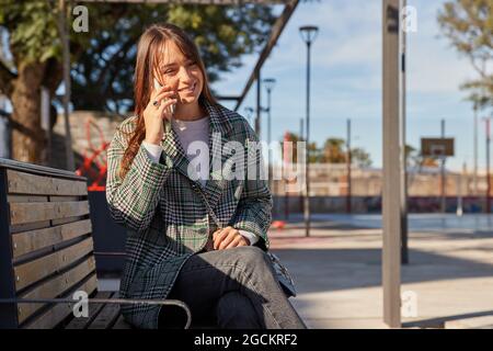 Moderne, lächelnde Millennial-Frau im stilvollen Frühlingsoutfit, die auf der Bank sitzt und Anrufe entgegennimmt, während sie sich auf der städtischen Straße ausruht und wegschaut Stockfoto