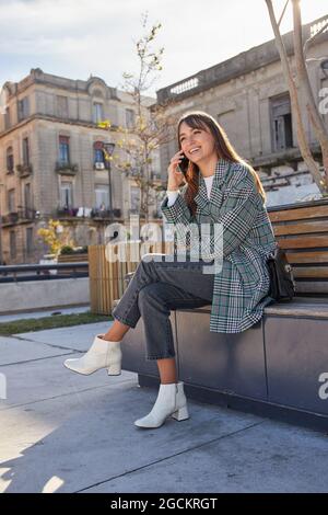 Die gesamte Länge der modernen Millennial-Frau in stilvollem Frühlingsoutfit sitzt auf der Bank und nimmt Telefonanruf ab, während sie sich auf der städtischen Straße ausruht Stockfoto