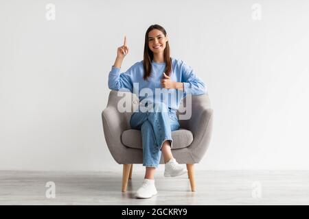 Fröhliche junge Dame zeigt nach oben und zeigt den Daumen nach oben, sitzt im Sessel an der weißen Studiowand Stockfoto