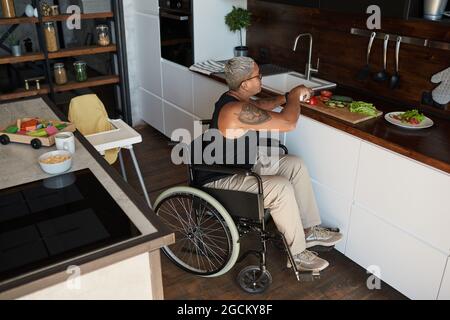 Hochwinkelporträt einer zeitgenössischen tätowierten Frau im Rollstuhl, die Salat zu Hause in der Küche kocht, Kopierraum Stockfoto