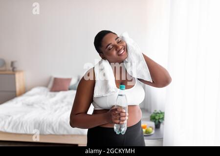 Plus size schwarze Frau mit einer Flasche Wasser wischen Schweiß mit Handtuch nach dem inländischen Training, drinnen Stockfoto