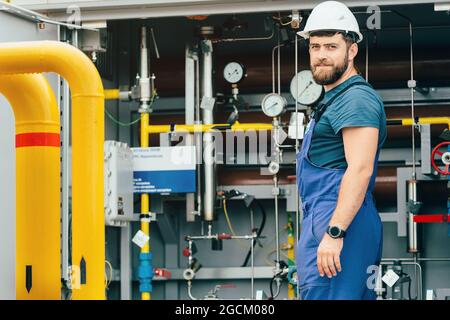 Porträt eines Gasarbeiters in einem Helm und einem Arbeitsüberfall vor dem Hintergrund von Geräten mit Rohren und Manometern. Gasodoriereinheit. Betreiber der Gasverteilerstation. Stockfoto