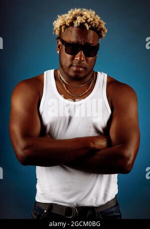 Dies ist ein Studioporträt eines Afrikaners in Sonnenbrillen und einem weißen T-Shirt auf dunkelblauem Hintergrund. Ein schwül getragener Kerl mit modischer Frisur und Zopf hält seine Hände auf der Brust und blickt direkt in die Kamera. Stockfoto