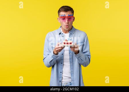 Feier, Urlaub und Menschen Emotionen Konzept. Verträumt glücklich schöner Mann machen Wunsch auf Geburtstagskuchen, mit Party, tragen lustige Sonnenbrille Stockfoto