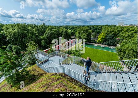 London, Großbritannien. 9 August 2021. Der Blick von oben nach Westen, wo die Baustelle durch einen frischen Rasen ersetzt wurde - den £2 Million Marble Arch Mound - wurde im Februar angekündigt und wurde gerade im August nach einem „Fehlstart“ kostenlos wieder eröffnet. Besucher können auf den 25 Meter hohen Gipfel klettern, von dem aus man von einer Aussichtsplattform aus einen 360-Grad-Blick auf die Oxford Street und den Hyde Park hat und von der breiten Öffentlichkeit nie zuvor gesehene Ausblicke genießen kann. Auf dem Weg nach unten steigen die Besucher in einen ausgehöhlten Raum, der für Ausstellungen genutzt wird. Es ist bis zum 2022. Januar für Besucher geöffnet.“ Entworfen von der niederländischen Firma Stockfoto