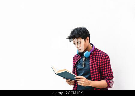 Asiatischer Hipster junger Mann, der ein Buch gegen eine weiße Wand liest - asiatischer Student, der ein Buch in den Händen hält und dies während der Freizeit im Freien liest - Le Stockfoto
