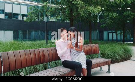Kranschuss des glücklichen Mixed-Race-Paares mit Datum in einem schönen Stadtpark. Sie sitzen auf der Bank, lächeln und reden miteinander. Stockfoto