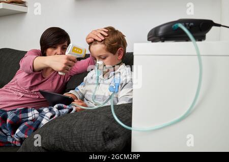 Liebevolle Mutter mit Infrarot-Thermometer Messung der Temperatur des Jungen Atmung in Sauerstoffmaske während Inhalationsverfahren und beobachten Cartoon auf Tabelle Stockfoto