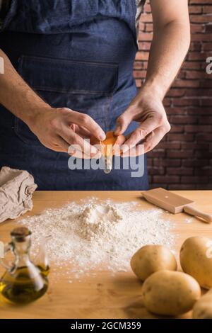 Nicht erkennbarer Mann in der Schürze, der während der Zubereitung des Gnocchi-Teigs auf dem Tisch in der Küche in der Nähe von Kartoffeln und Öl Eigelb in einen Haufen Weizenmehl zerbrach Stockfoto