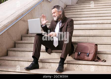 Positiv gealterter bärtiger Mann in elegantem Anzug, der auf einer Treppe sitzt und mit der Hand winkt und beim Online-Videotreffen über einen Laptop Hallo sagt Stockfoto