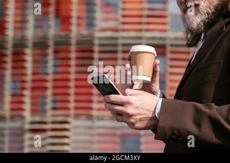 Seitenansicht des Getreides nicht erkennbarer grauhaariger bärtiger Mann im formellen Anzug, der Kaffee zum Mitnehmen trinkt und auf der Stadtstraße das Mobiltelefon durchstöbert Stockfoto