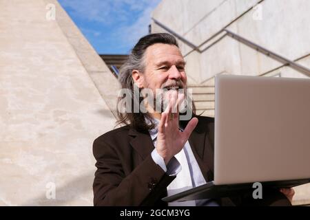 Von unten positiv gealterter bärtiger Mann in elegantem Anzug, der auf einer Treppe sitzt und mit der Hand winkt und beim Online-Videotreffen über einen Laptop Hallo sagt Stockfoto