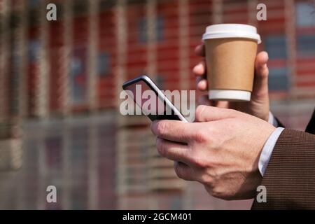 Seitenansicht des Getreides nicht erkennbarer grauhaariger bärtiger Mann im formellen Anzug, der Kaffee zum Mitnehmen trinkt und auf der Stadtstraße das Mobiltelefon durchstöbert Stockfoto