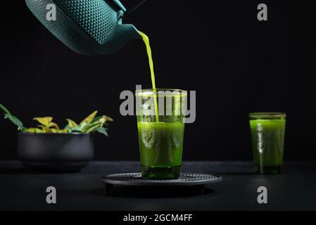 Gesunder japanischer Matcha-Tee, der während der Teezeremonie vor schwarzem Hintergrund aus grüner Teekannen in Glas gegossen wird Stockfoto